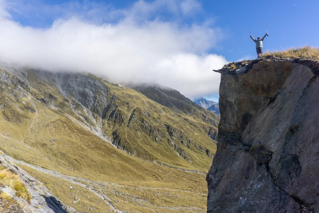 Woman standing close to the edge of a cliff with arms up, above the Rees Saddle on the Rees Dart Track
