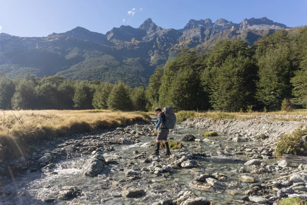 Woman with a large packing crossing a small stream with mountains in the background